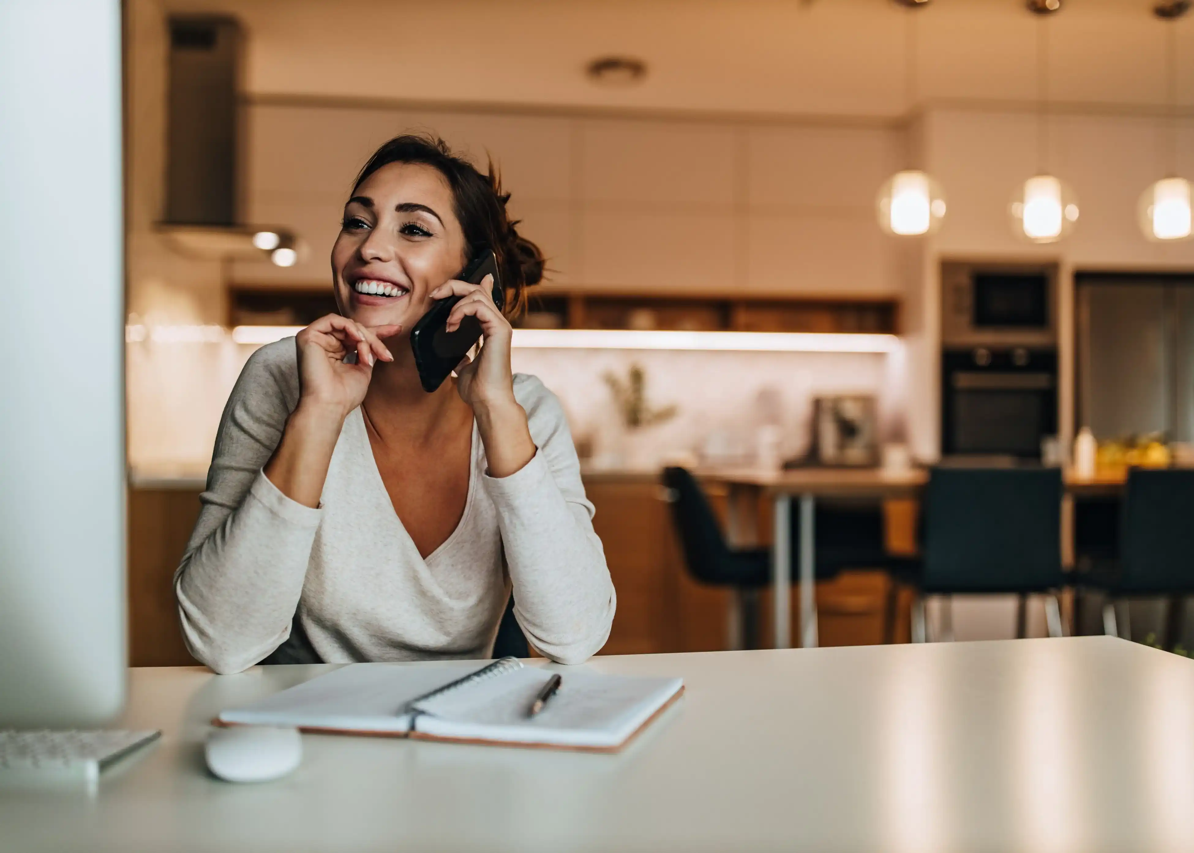 Smiling woman talking on a phone while sitting at a desk with an open notebook in a modern kitchen.