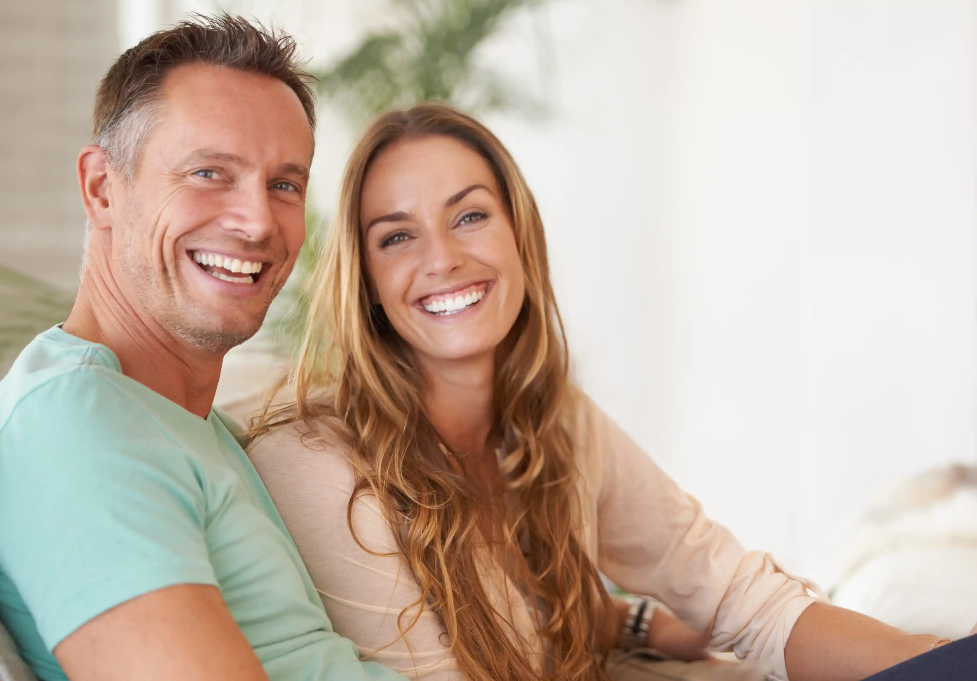 Smiling middle-aged man in turquoise shirt and woman with long hair in beige top sitting closely together indoors.